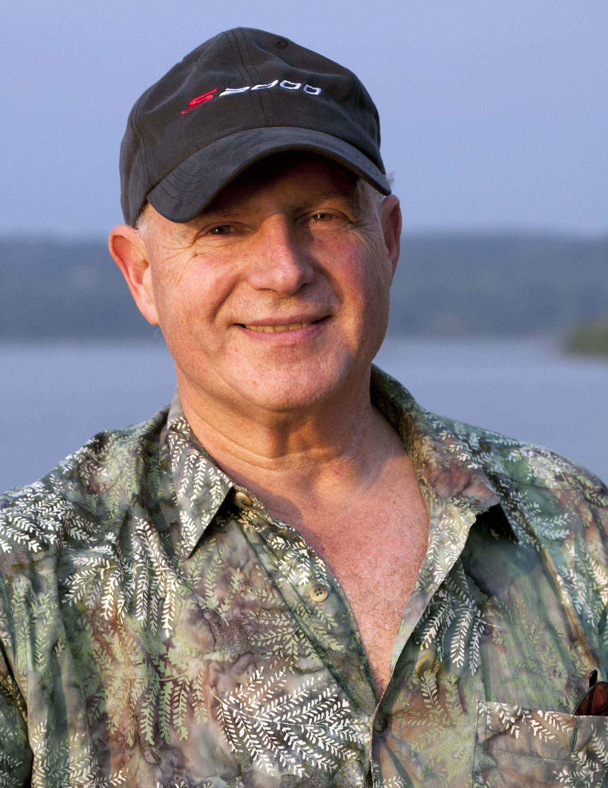 Headshot of elder man wearing a shirt and hat outdoors against a lake or mountain background.